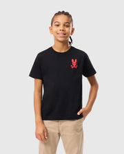 A young person with braided hair smiles while wearing a stylish Psycho Bunny KIDS NEW YORK CITY TEE in black, crafted from soft Pima cotton and featuring a red emblem of a bunny and crossbones. They pair it with beige pants, one hand casually in their pocket, standing against a plain white background.