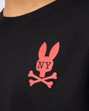 Close-up of a black Psycho Bunny KIDS NEW YORK CITY TEE - B0U554W1PC, crafted from premium Pima cotton and featuring a red logo with a stylized rabbit head above crossed bones. The letters "NY" are printed on the rabbit's face, making this tee both stylish and comfortable for kids.