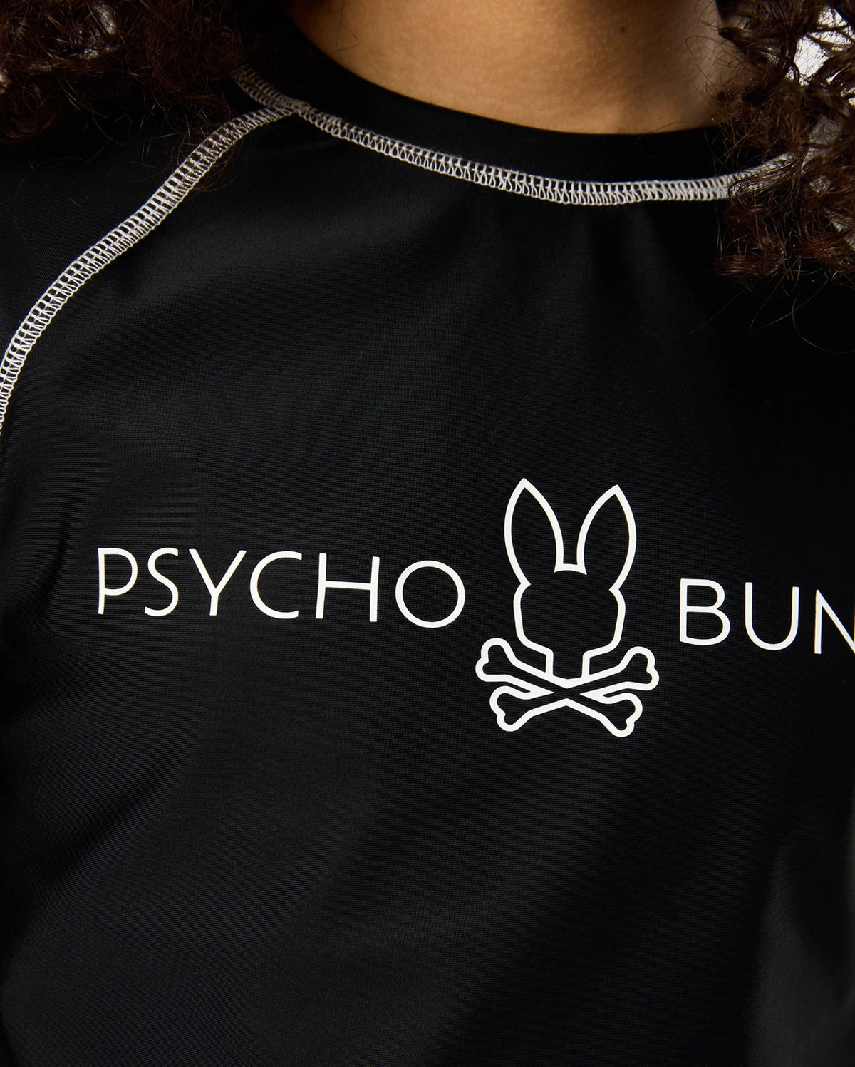 Close-up of a person wearing a black shirt with white seam detailing. The KIDS SHORT SLEEVE ANTI-UV RASHGUARD - B0U558C200 by Psycho Bunny features the words "Psycho Bunny" and a logo of a bunny head with crossbones underneath, promising UPF 50+ protection for ultimate safety in the sun.