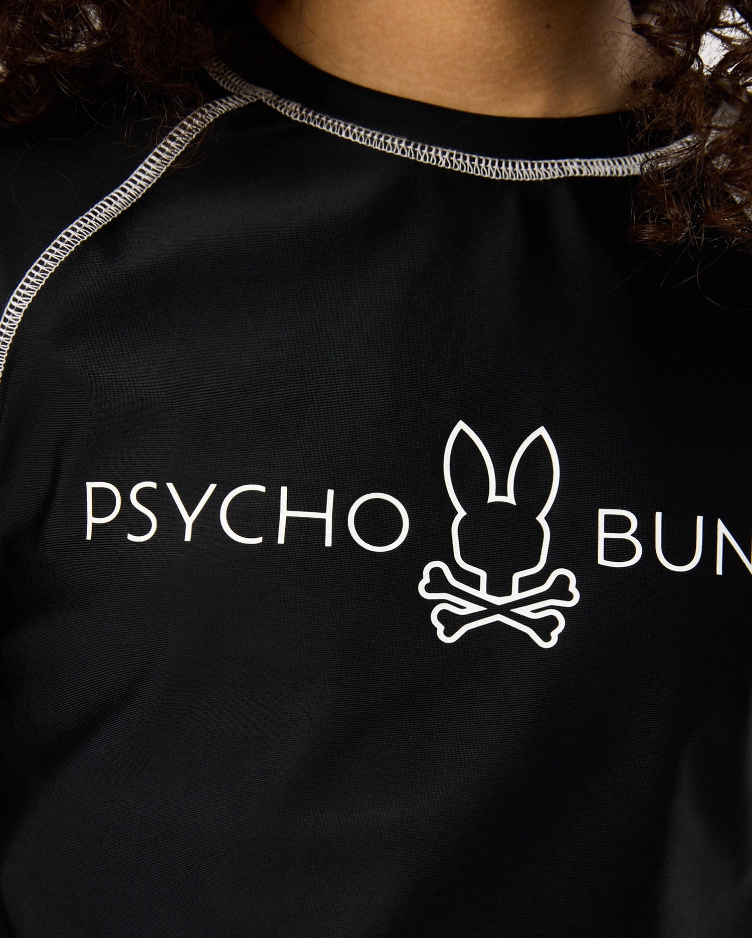 Close-up of a person wearing a black shirt with white seam detailing. The KIDS SHORT SLEEVE ANTI-UV RASHGUARD - B0U558C200 by Psycho Bunny features the words "Psycho Bunny" and a logo of a bunny head with crossbones underneath, promising UPF 50+ protection for ultimate safety in the sun.