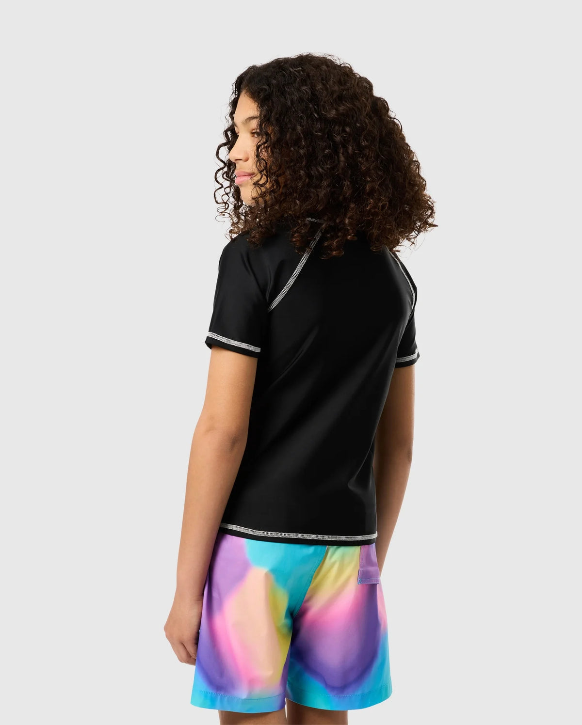 A person with curly hair stands facing slightly away, wearing a black short-sleeved shirt with white seam details and colorful tie-dye shorts in shades of blue, pink, and yellow. The UPF 50+ protection of the Psycho Bunny Kids Short Sleeve Anti-UV Rashguard - B0U558C200 ensures they're ready for the sun. The background is plain and light-colored.