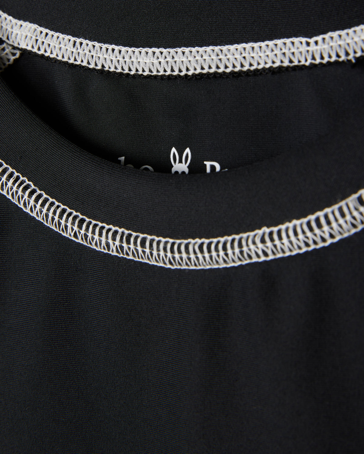 Close-up image of a black KIDS SHORT SLEEVE ANTI-UV RASHGUARD - B0U558C200 with white stitching around the neckline of the garment. The inside label is partially visible, featuring a small white outline of a rabbit head. This short sleeve rashguard offers UPF 50+ protection for ultimate sun safety from Psycho Bunny.