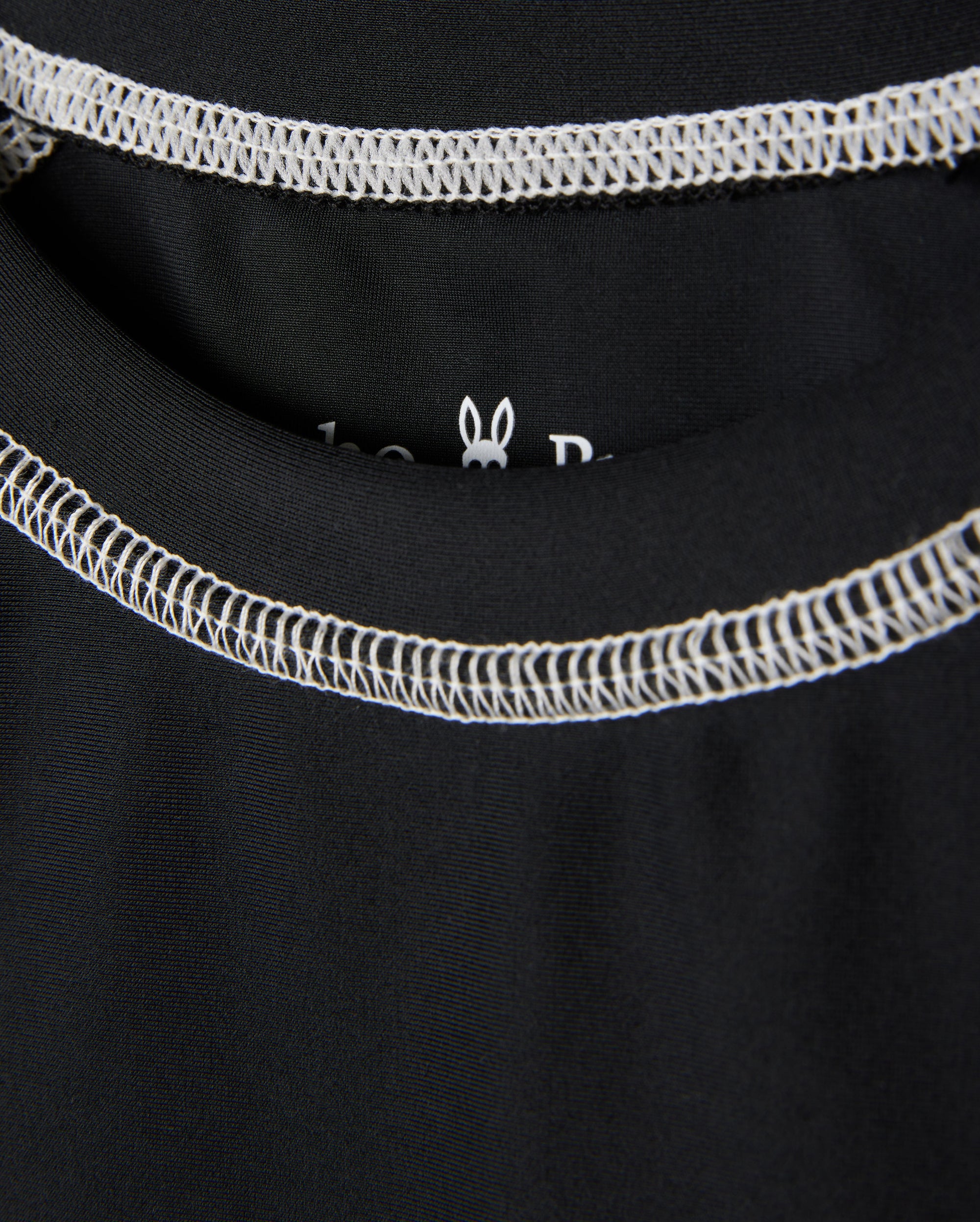 Close-up image of a black KIDS SHORT SLEEVE ANTI-UV RASHGUARD - B0U558C200 with white stitching around the neckline of the garment. The inside label is partially visible, featuring a small white outline of a rabbit head. This short sleeve rashguard offers UPF 50+ protection for ultimate sun safety from Psycho Bunny.
