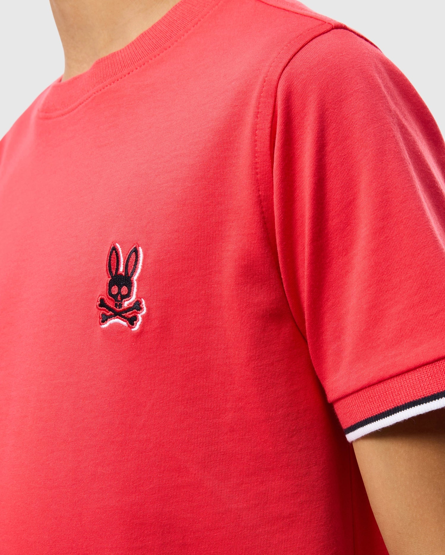 Close-up of a person wearing a red Psycho Bunny KIDS KAYDEN FASHION TEE - B0U577C200 with an embroidered chest Bunny skull and crossbones logo. The sleeve has a white, black, and red striped cuff detail. Made from soft Pima cotton, the background is plain and light-colored.