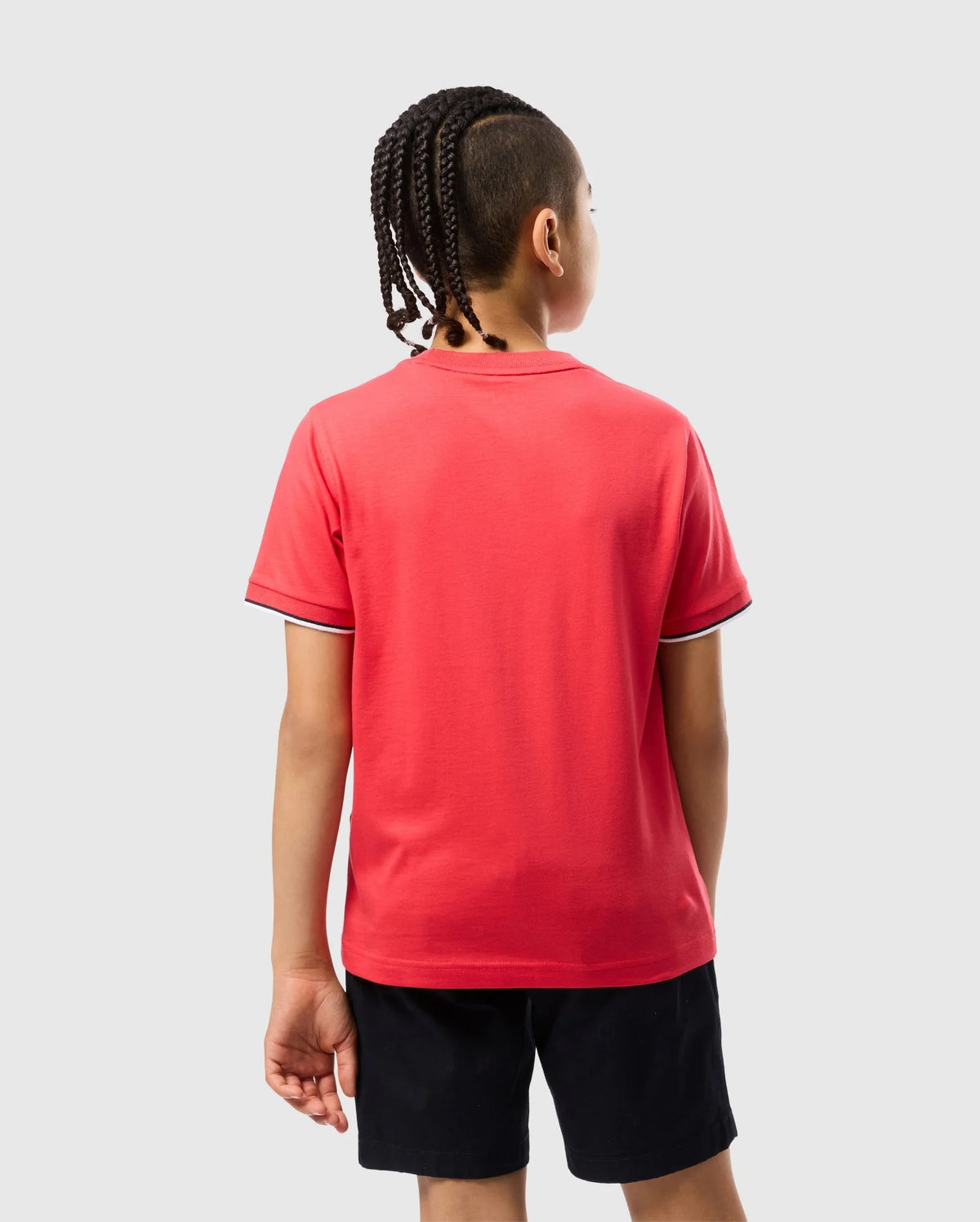 A person with braided hair is seen from the back, wearing a red short-sleeve Psycho Bunny KIDS KAYDEN FASHION TEE - B0U577C200 with white trim and black shorts. The background is plain and light gray.