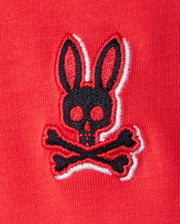 A close-up of an embroidered chest Bunny logo on red Pima cotton fabric. The logo features a black bunny head with long ears and large round eyes, positioned above a pair of crossed bones, reminiscent of a skull and crossbones design. The edges of the logo are trimmed in white, perfect for the KIDS KAYDEN FASHION TEE - B0U577C200 by Psycho Bunny.