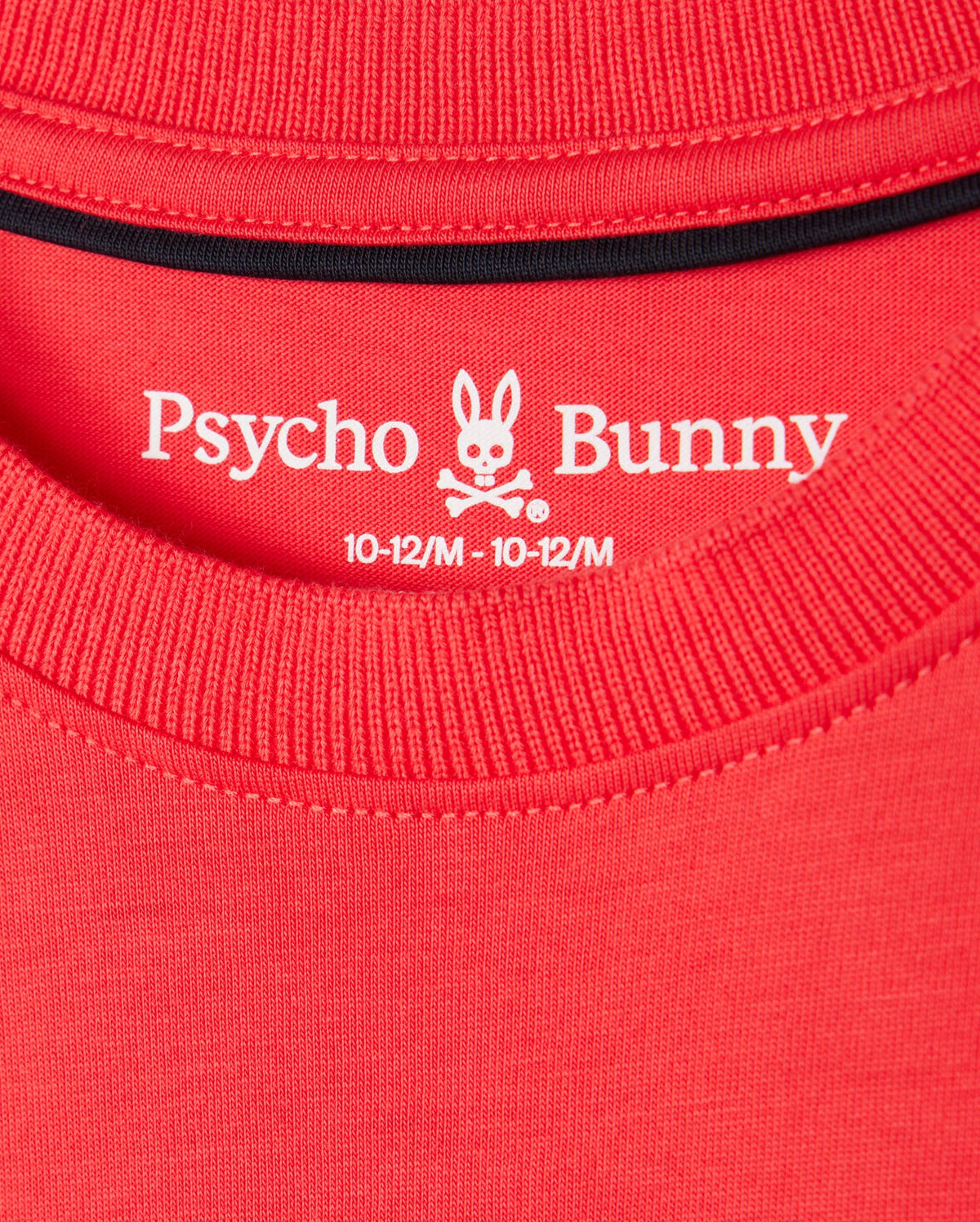 Close-up of the neck label on a red KIDS KAYDEN FASHION TEE - B0U577C200 by Psycho Bunny, featuring the brand's name and logo of a bunny skull with crossbones. The label indicates the size as 10-12/M. The stitching details of the collar and neckline are visible, highlighting the premium Pima cotton used for this stylish piece.