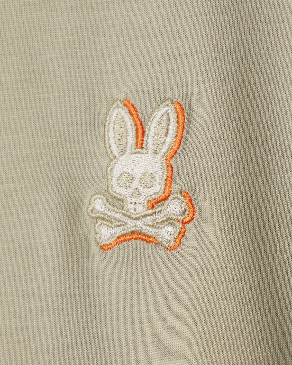 Close-up of a beige Pima cotton fabric with an embroidered chest logo featuring a stylized skull and crossbones, with the skull having bunny ears and highlighted with an orange outline. Perfect for a Psycho Bunny KIDS KAYDEN FASHION TEE - B0U577C200.