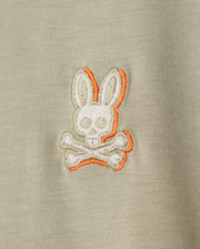 Close-up of a beige Pima cotton fabric with an embroidered chest logo featuring a stylized skull and crossbones, with the skull having bunny ears and highlighted with an orange outline. Perfect for a Psycho Bunny KIDS KAYDEN FASHION TEE - B0U577C200.