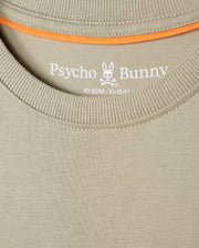 Close-up of a beige Psycho Bunny KIDS KAYDEN FASHION TEE - B0U577C200 featuring the brand's embroidered chest Bunny logo with crossbones, along with the size designation "10-12/M" printed on the inner collar area. Orange trim is visible along the inside of the collar. Crafted from soft Pima cotton for ultimate comfort.