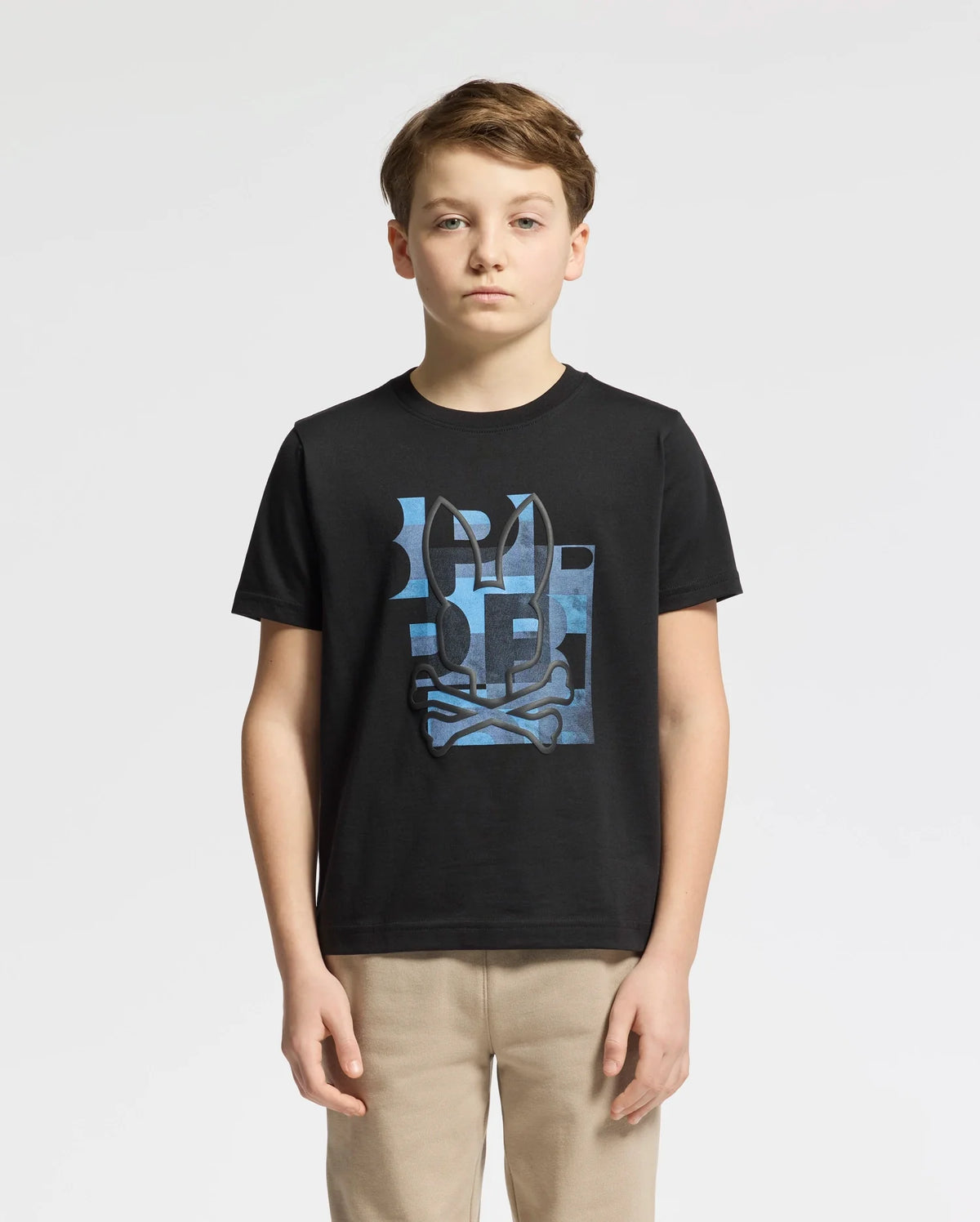 A boy stands against a plain background wearing the Psycho Bunny KIDS ZEALAND GRAPHIC TEE - B0U604F200 in black, made from soft Pima cotton, with beige pants. The shirt features a stylized blue and white pattern with abstract elements and a playful bunny logo.