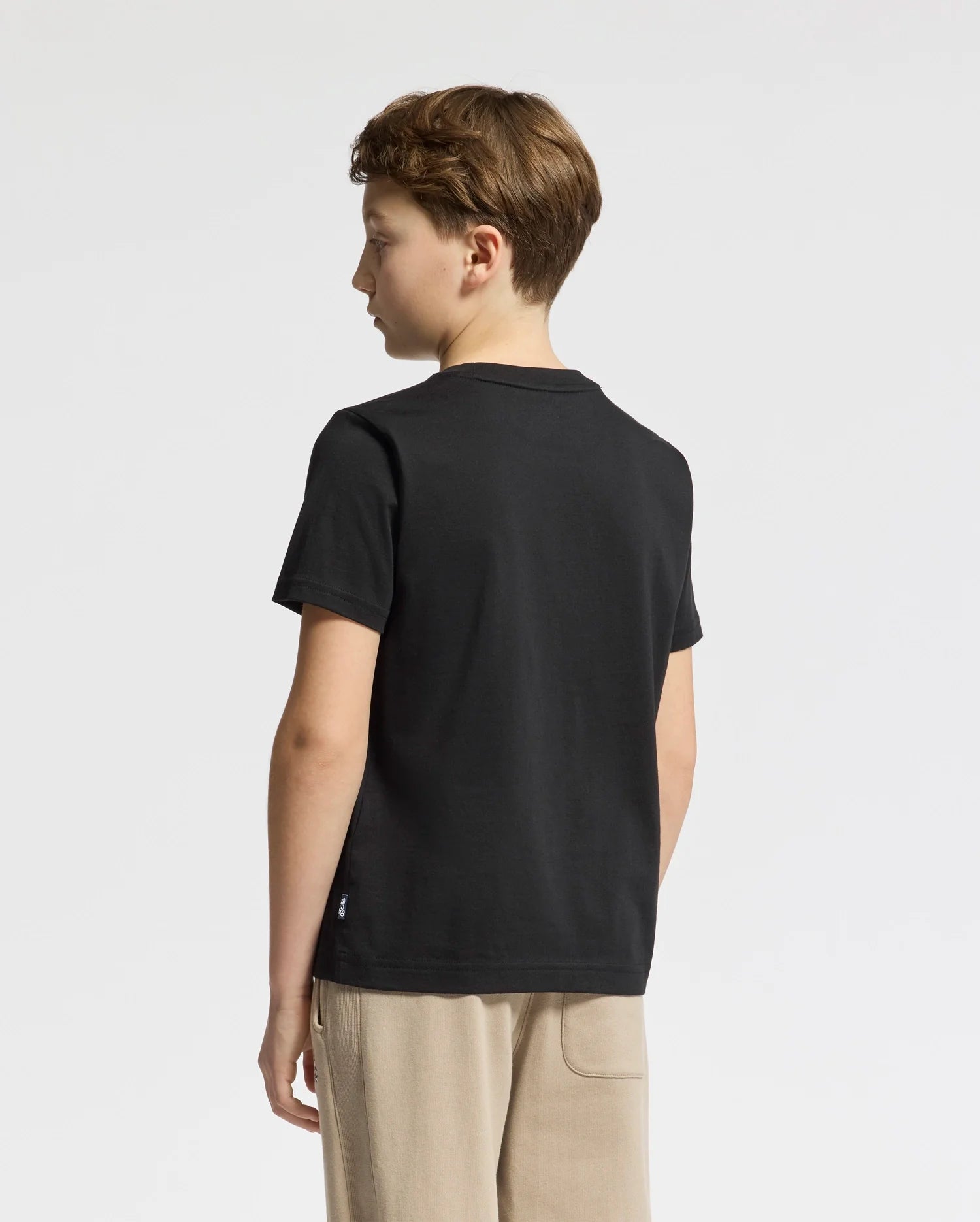 A young boy with brown hair stands sideways, wearing beige pants and the Psycho Bunny KIDS ZEALAND GRAPHIC TEE (B0U604F200), made from soft Pima cotton. The black T-shirt features a subtle bunny logo, and he gazes slightly downward with a neutral expression against the plain white background.