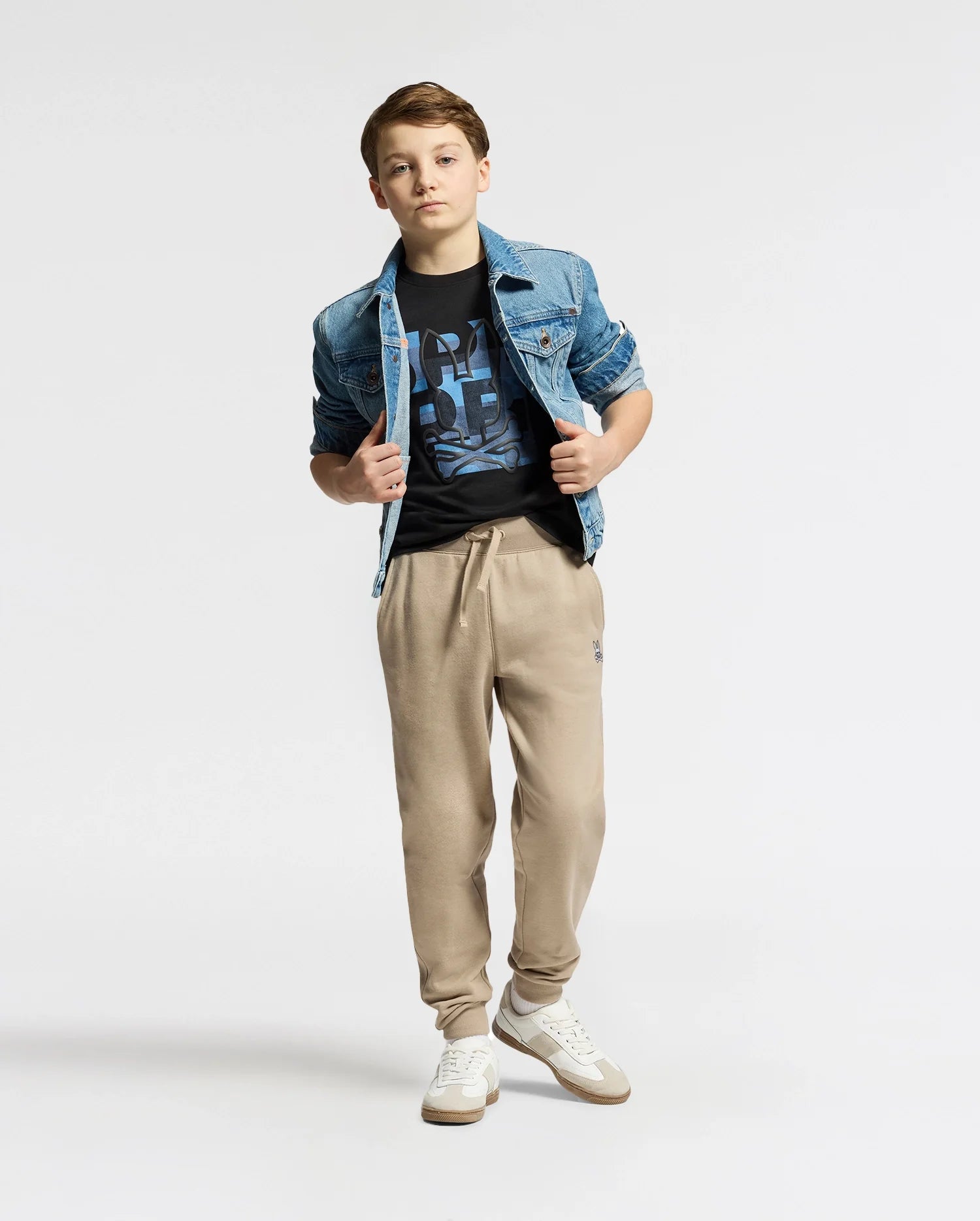 A young person stands wearing a denim jacket over the KIDS ZEALAND GRAPHIC TEE by Psycho Bunny, beige jogger pants, and white sneakers. They pose with hands in pockets against a plain white backdrop.