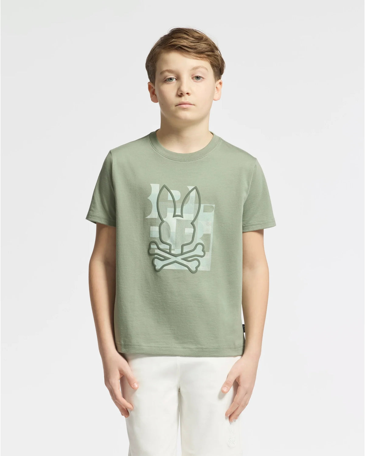 A boy with short light brown hair stands against a plain white background, wearing the KIDS ZEALAND GRAPHIC TEE - B0U604F200 by Psycho Bunny. This soft Pima cotton tee displays a big bunny logo with bones on the front and is paired with crisp white pants, matching his neutral expression.