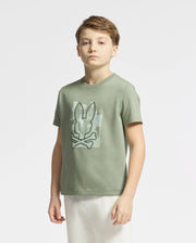 A young boy with light brown hair wears a green Psycho Bunny KIDS ZEALAND GRAPHIC TEE - B0U604F200, crafted from soft Pima cotton and showcasing a bunny and crossbones design. He stands against a plain white background, gazing forward with a neutral expression.