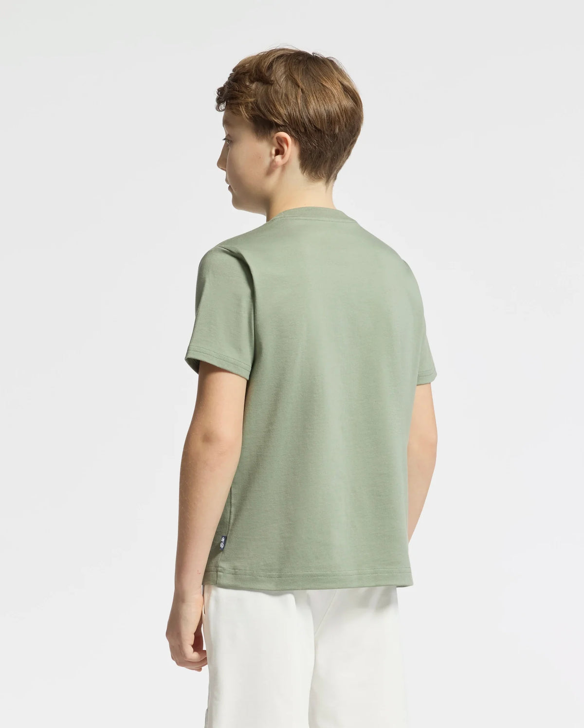 A boy is facing away from the camera, wearing a Psycho Bunny KIDS ZEALAND GRAPHIC TEE - B0U604F200 in light green with a large bunny logo and white shorts. He stands against a white background.