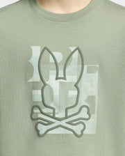 Close-up of a person wearing a Psycho Bunny KIDS ZEALAND GRAPHIC TEE - B0U604F200 in green Pima cotton, featuring a large raised bunny logo with crossbones design in a playful tone.