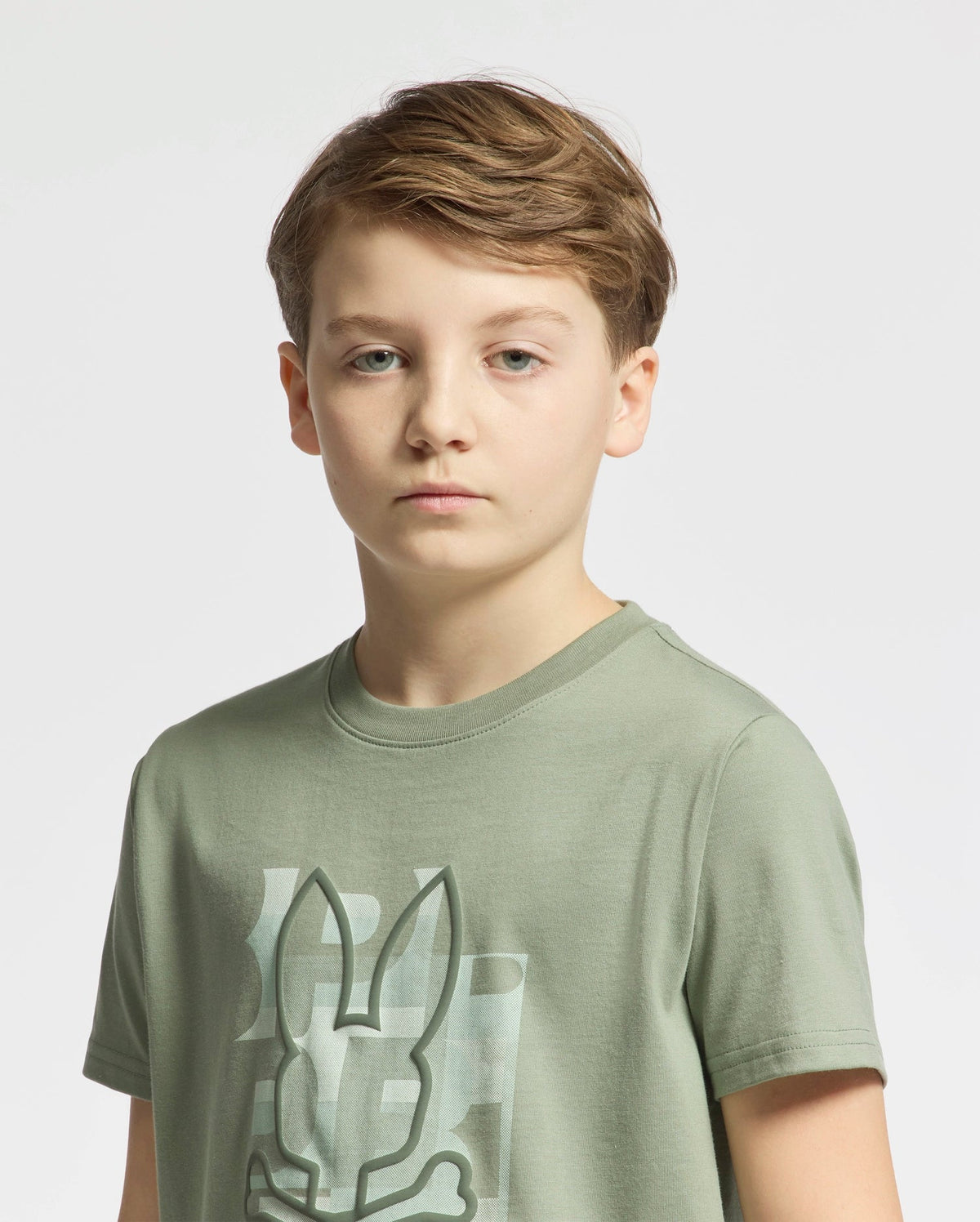 A young boy with short light brown hair stands against a plain background, wearing a Psycho Bunny KIDS ZEALAND GRAPHIC TEE in light green, featuring a prominent bunny logo. He has a neutral expression and looks directly at the camera.