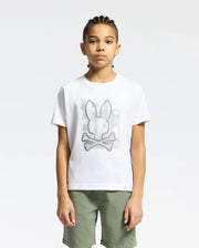 A young person is sporting a Psycho Bunny KIDS ZEALAND GRAPHIC TEE - B0U604F200 featuring a bunny with crossbones. Made from soft Pima cotton, the shirt complements their braided hair and light green shorts against a plain white backdrop.
