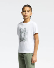 A young person with braided hair stands against a light background, wearing the white KIDS ZEALAND GRAPHIC TEE by Psycho Bunny. Crafted from soft Pima cotton, it features a big bunny skull logo and pairs perfectly with green pants.