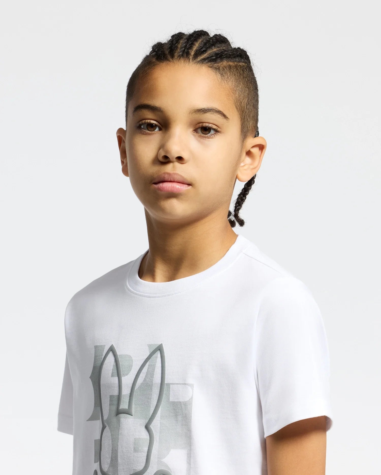 A young person with braided hair wears the Psycho Bunny KIDS ZEALAND GRAPHIC TEE (B0U604F200), crafted from crisp Pima cotton with an abstract graphic, against a plain gray background.