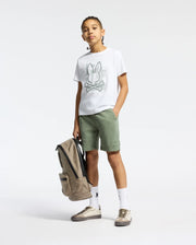 A young person stands confidently in a Psycho Bunny KIDS ZEALAND GRAPHIC TEE, green shorts, and sneakers, holding a beige backpack against a plain white background.