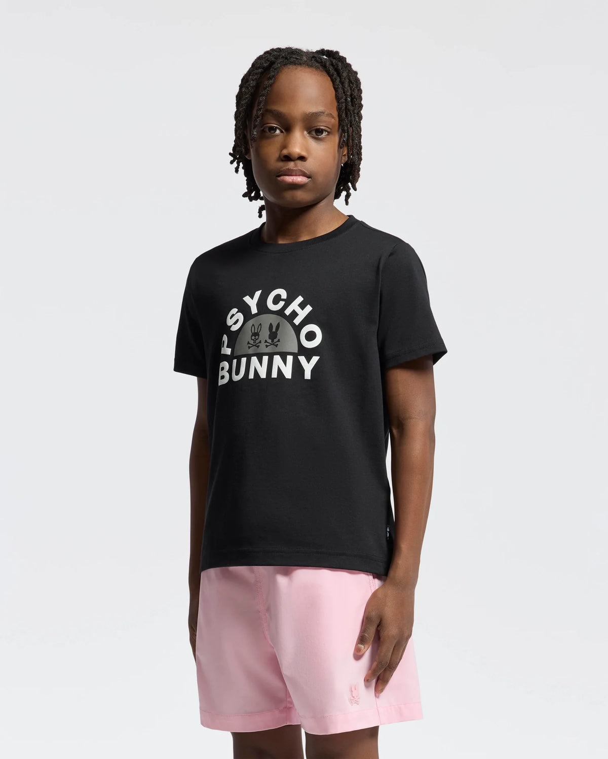 A young person with short dreadlocks wears a black Psycho Bunny KIDS GOLDIE GRAPHIC TEE, showcasing a white graphic and crafted from soft Peruvian Pima cotton. They pair it with pink shorts, standing against a plain white background, facing forward.