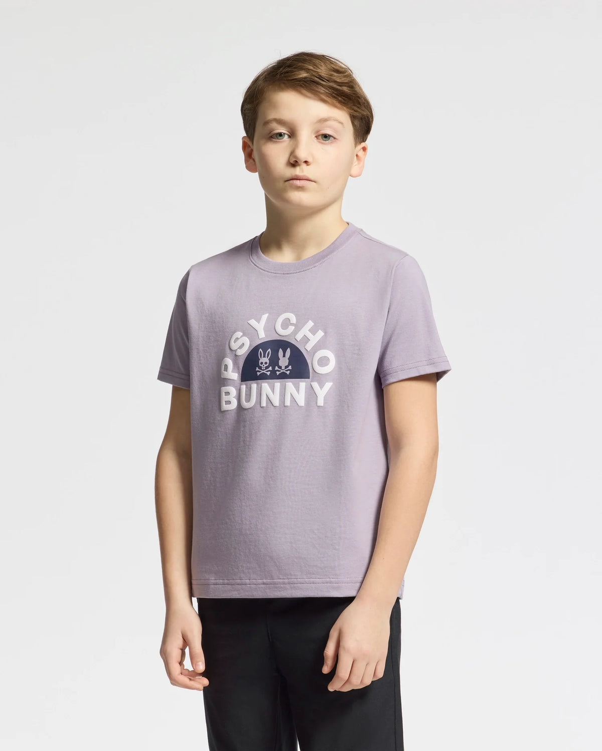 A young person stands against a plain backdrop, wearing the Kids Goldie Graphic Tee (B0U606F200) in light purple by Psycho Bunny. Made from soft Peruvian Pima cotton, the tee matches their short, light brown hair as they gaze at the camera with a neutral expression.