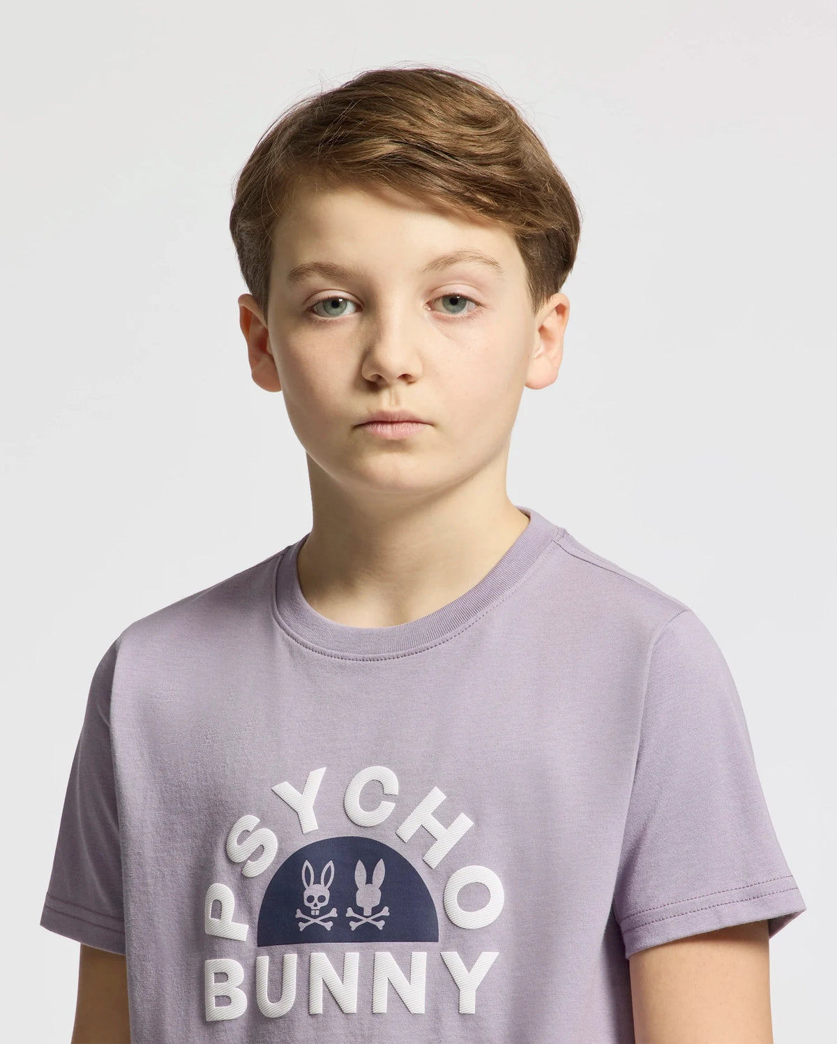 The boy with short hair stands against a plain background, wearing the stylish lavender KIDS GOLDIE GRAPHIC TEE by Psycho Bunny (B0U606F200), crafted from luxurious Peruvian Pima cotton, featuring the iconic bunny and crossbones graphic.