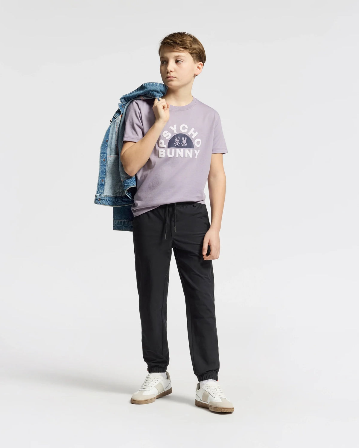 A boy poses against a plain backdrop, casually draping a denim jacket over his shoulder. He's wearing a lavender "Psycho Bunny" KIDS GOLDIE GRAPHIC TEE with a bunny design, black jogger pants, and white Peruvian Pima cotton sneakers. His thoughtful gaze is directed to the side.