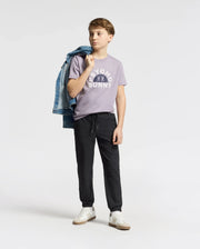 A boy poses against a plain backdrop, casually draping a denim jacket over his shoulder. He's wearing a lavender "Psycho Bunny" KIDS GOLDIE GRAPHIC TEE with a bunny design, black jogger pants, and white Peruvian Pima cotton sneakers. His thoughtful gaze is directed to the side.