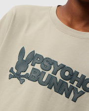 Close-up of a child wearing the KIDS MELVIN GRAPHIC TEE by Psycho Bunny, in beige Pima cotton. The t-shirt showcases a stylized graphic design of a bunny head with crossbones and the text "PSYCHO BUNNY" in textured dark print. The wordmark graphic is rendered in an HD-silicone print on a plain background, focusing on the chest area.