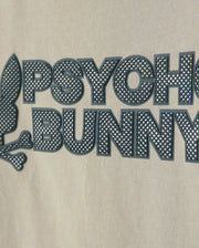 Close-up of a beige fabric with a raised graphic design showcasing an HD-silicone print. The image features the words "PSYCHO BUNNY" in black, capital letters beside a stylized rabbit head and crossbones emblem. Crafted from premium Pima cotton, this is the KIDS MELVIN GRAPHIC TEE - B0U663C200 by Psycho Bunny.