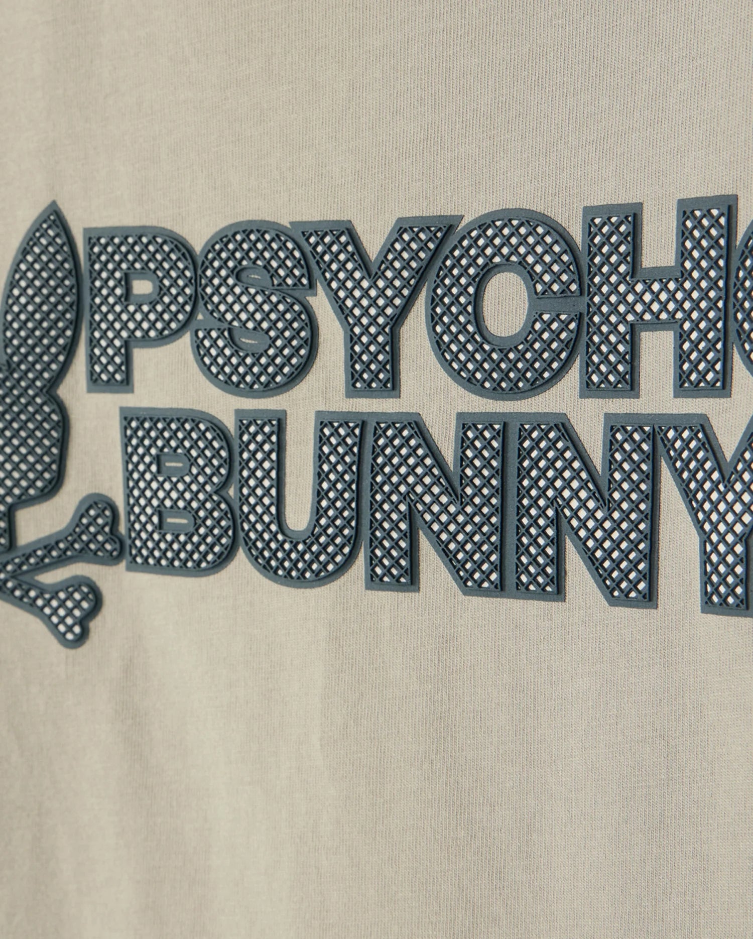 Close-up of a beige fabric with a raised graphic design showcasing an HD-silicone print. The image features the words "PSYCHO BUNNY" in black, capital letters beside a stylized rabbit head and crossbones emblem. Crafted from premium Pima cotton, this is the KIDS MELVIN GRAPHIC TEE - B0U663C200 by Psycho Bunny.