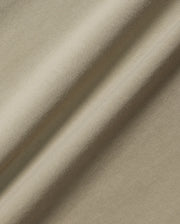 A close-up of the beige Pima cotton fabric of the KIDS MELVIN GRAPHIC TEE - B0U663C200 from Psycho Bunny, showcasing its smooth texture and subtle diagonal folds. The lighting highlights the gentle sheen and soft appearance of this textile.