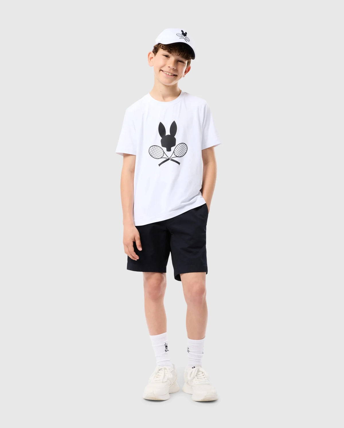 A young boy is standing against a plain background, smiling with his hands in his pockets. He is wearing a white KIDS COURTSIDE GRAPHIC TEE - B0U681C200 from Psycho Bunny featuring a bunny and crossed tennis racquets, black shorts, white socks, white sneakers, and a white cap with the same bunny logo. The look is tennis-inspired and perfect for an active day.