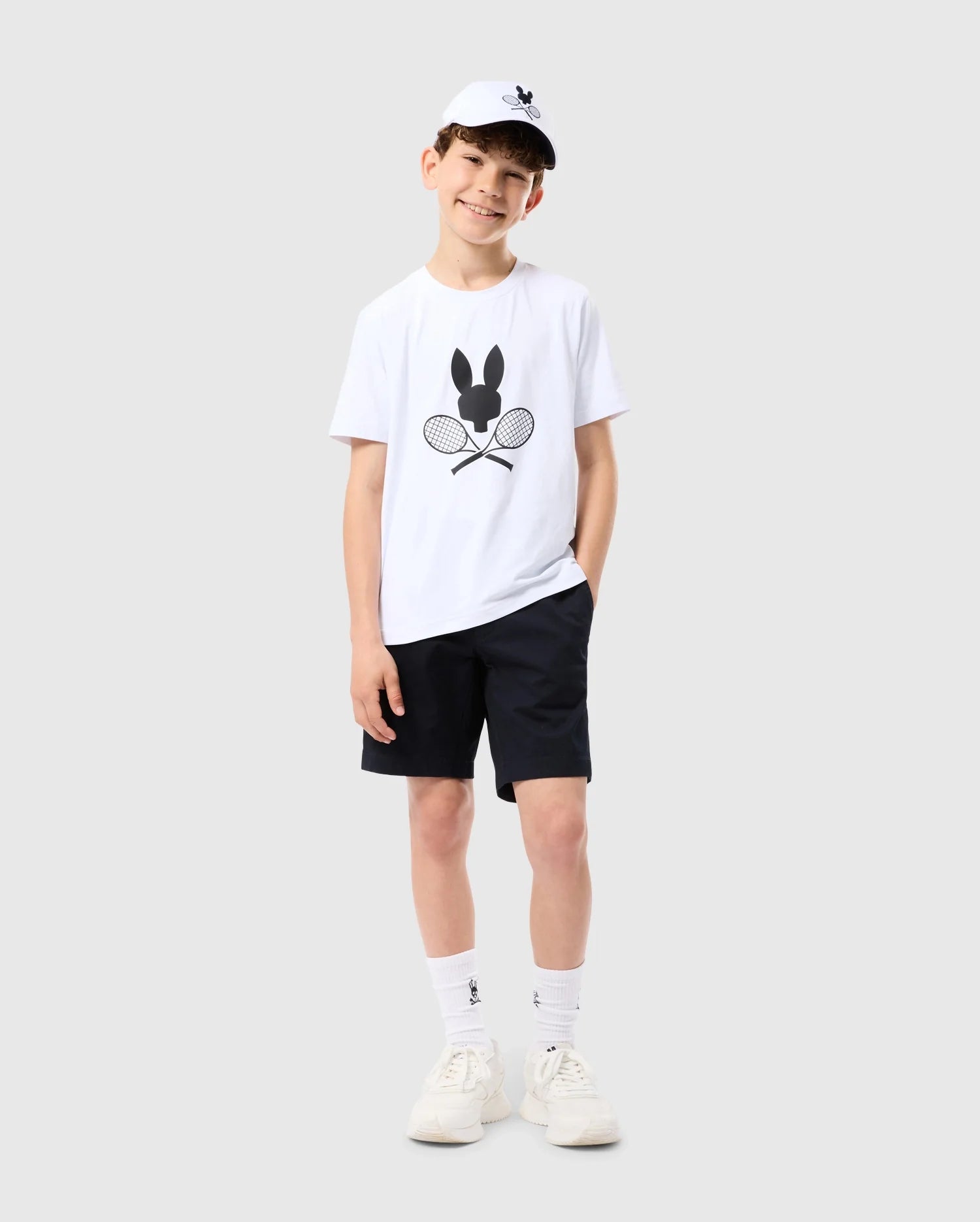 A young boy is standing against a plain background, smiling with his hands in his pockets. He is wearing a white KIDS COURTSIDE GRAPHIC TEE - B0U681C200 from Psycho Bunny featuring a bunny and crossed tennis racquets, black shorts, white socks, white sneakers, and a white cap with the same bunny logo. The look is tennis-inspired and perfect for an active day.