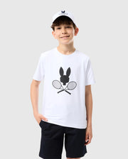 A smiling boy is standing against a plain light gray background. He wears a Psycho Bunny KIDS COURTSIDE GRAPHIC TEE - B0U681C200 featuring a black bunny and crossed tennis racket design, black shorts, and a white baseball cap. With his left hand in his pocket, he shows off his tennis-inspired look, complete with short, curly brown hair.
