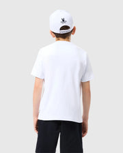 A young boy is standing with his back to the camera. He is wearing a white, KIDS COURTSIDE GRAPHIC TEE - B0U681C200 by Psycho Bunny, black shorts, and a white cap with a black graphic on the back. The background is plain, light grey.
