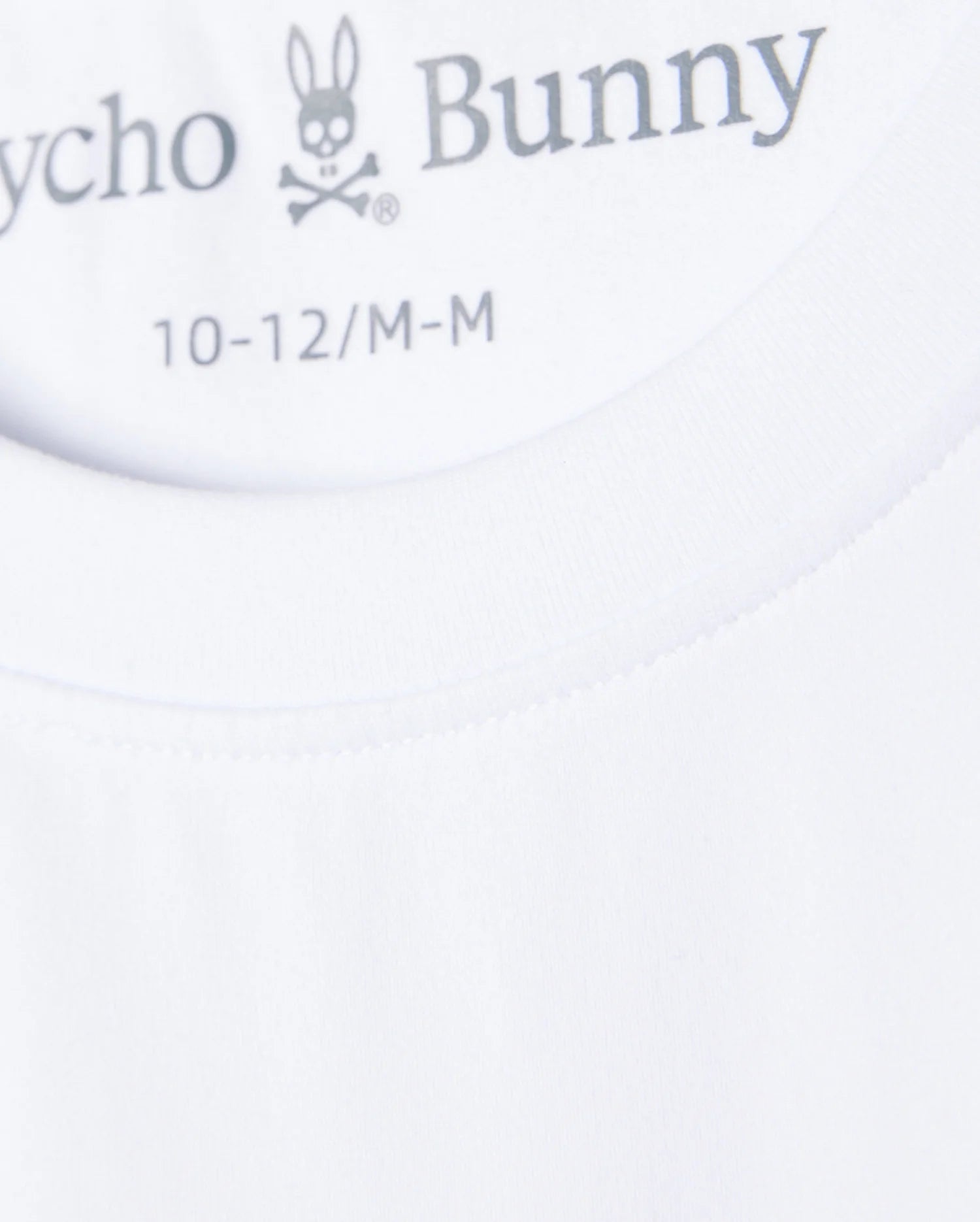 Close-up image of the neck tag of a white Psycho Bunny KIDS COURTSIDE GRAPHIC TEE - B0U681C200, displaying the brand logo featuring a rabbit skull and crossbones. The sizes indicated are 10-12/M-M. The fabric appears smooth and well-stitched, perfect for a tennis-inspired look.