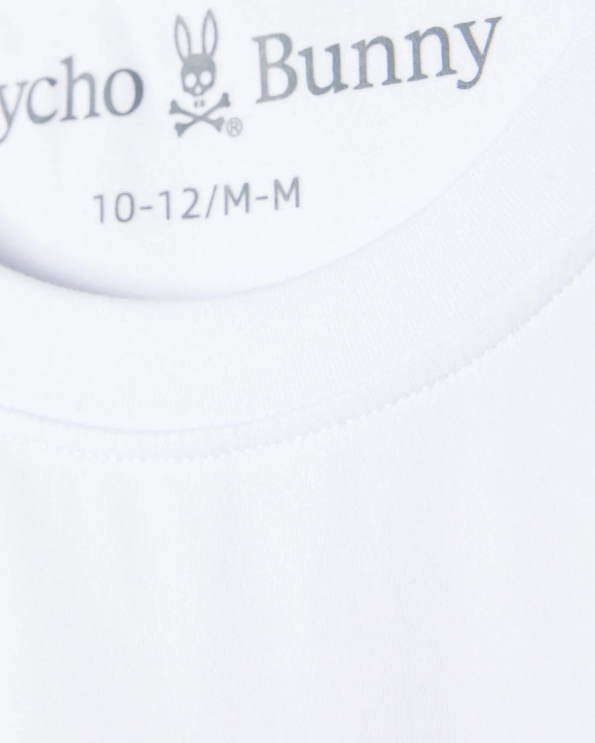 Close-up image of the neck tag of a white Psycho Bunny KIDS COURTSIDE GRAPHIC TEE - B0U681C200, displaying the brand logo featuring a rabbit skull and crossbones. The sizes indicated are 10-12/M-M. The fabric appears smooth and well-stitched, perfect for a tennis-inspired look.