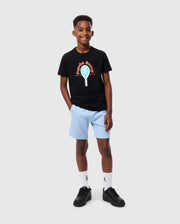 A young boy stands smiling, dressed in the Psycho Bunny KIDS COURTSIDE GRAPHIC TEE - B0U682C200, which showcases a vibrant design and text. He pairs it with light blue shorts, white socks, and black shoes. With his left hand casually tucked in his pocket against a plain gray background, his ensemble exudes an effortlessly tennis-inspired vibe.