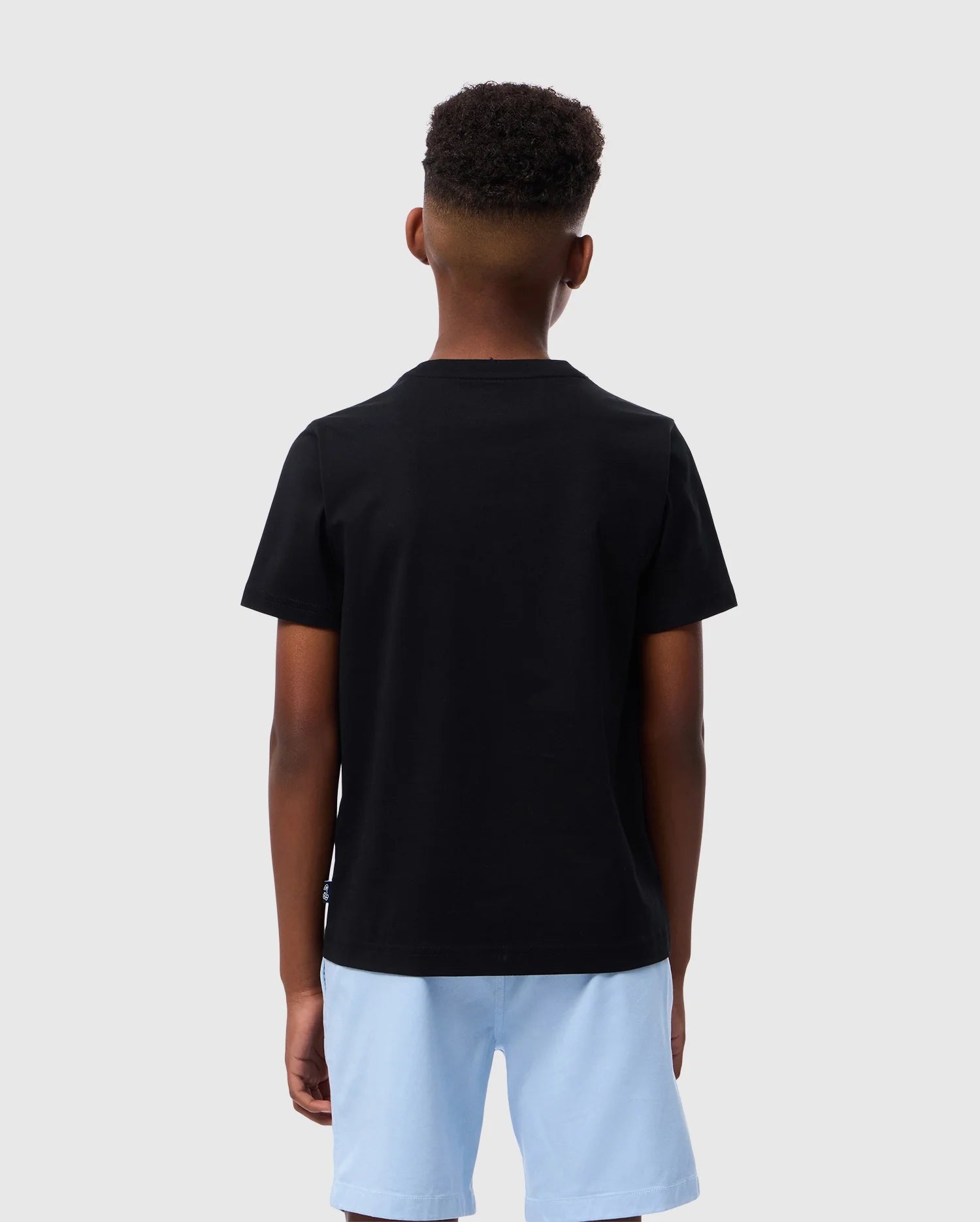 A young person with short curly hair is facing away, wearing the Psycho Bunny KIDS COURTSIDE GRAPHIC TEE - B0U682C200 in black and light blue shorts, reminiscent of a tennis-inspired style, against a plain grey background.