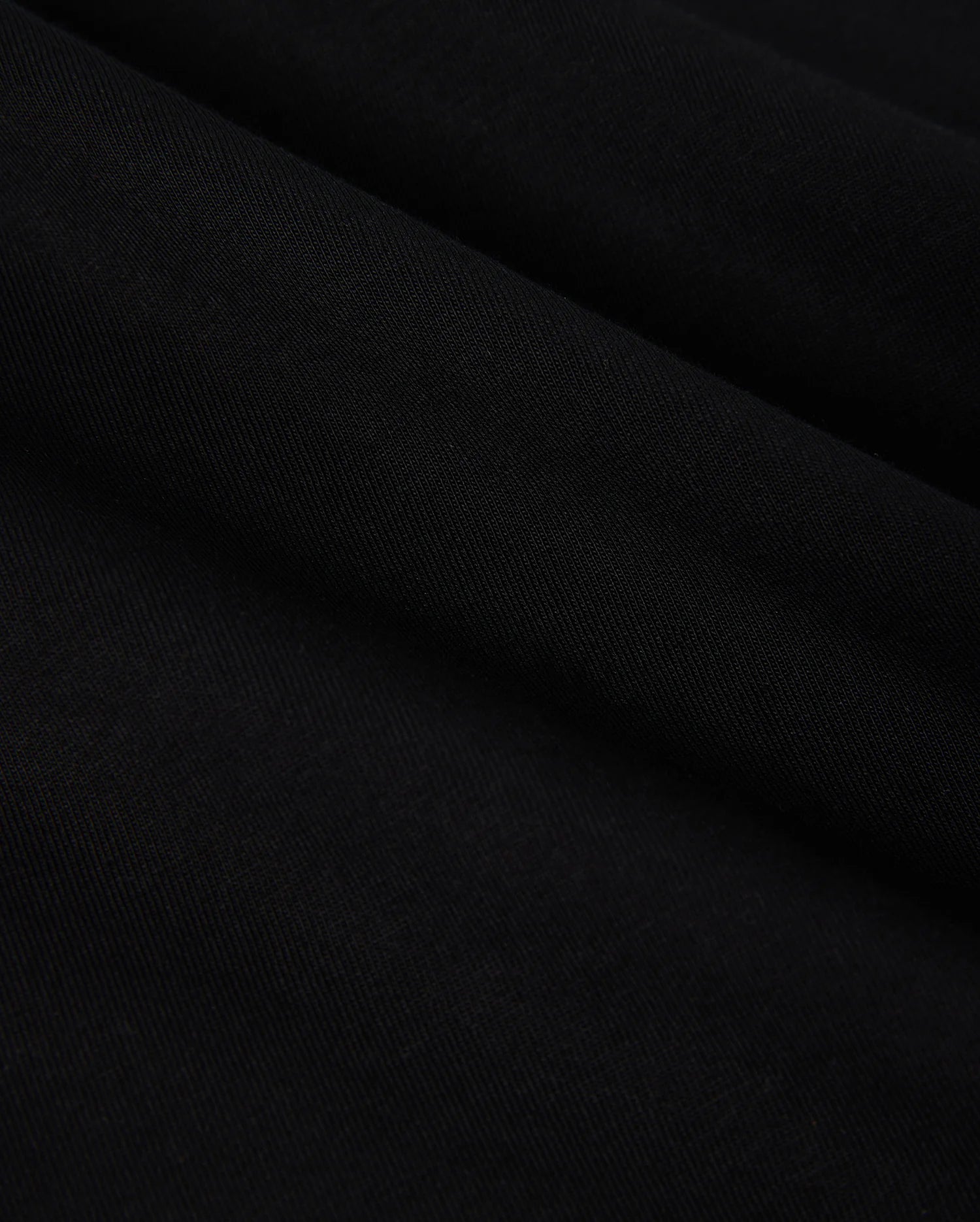 Close-up of the smooth, folded fabric of the KIDS COURTSIDE GRAPHIC TEE by Psycho Bunny in black, showcasing a subtle texture akin to a performance-led jersey. The material looks soft and slightly glossy, with gentle curves creating lines across the surface.