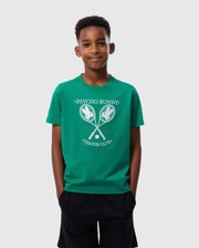 A young boy stands against a plain background wearing the Psycho Bunny KIDS COURTSIDE GRAPHIC TEE - B0U684C200 in green, featuring a "Psycho Bunny Tennis Club" print with two crossed tennis rackets. He has short, curly hair and dark shorts. He is smiling with his hands in his pockets, showcasing the stylish Pima cotton jersey.