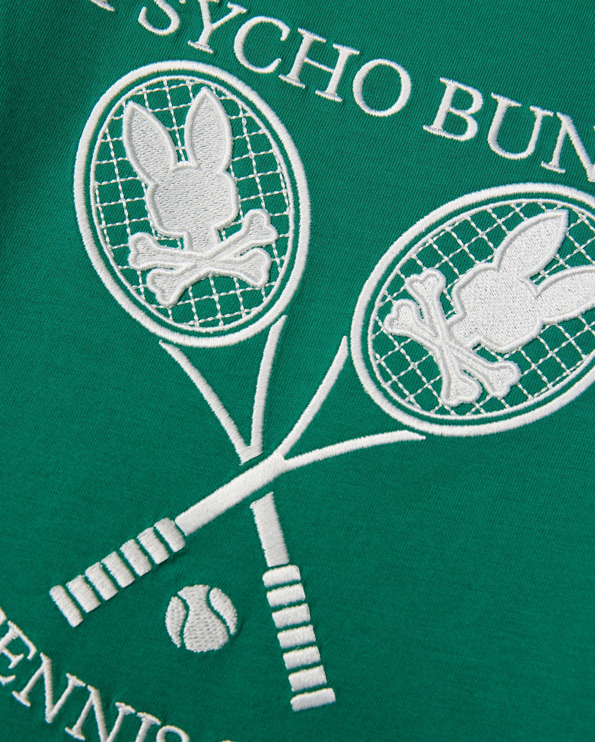 Close-up of a green Pima cotton jersey fabric with an embroidered white design from Psycho Bunny. The design on the Kids Courtside Graphic Tee - B0U684C200 features two intersecting tennis rackets with bunny skull-and-crossbones motifs in the racket heads and a small tennis ball below, perfect for a kids' tee. "PSYCHO BUNNY" and "TENNIS" are partially visible.
