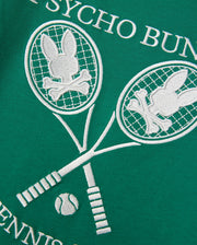 Close-up of a green Pima cotton jersey fabric with an embroidered white design from Psycho Bunny. The design on the Kids Courtside Graphic Tee - B0U684C200 features two intersecting tennis rackets with bunny skull-and-crossbones motifs in the racket heads and a small tennis ball below, perfect for a kids' tee. "PSYCHO BUNNY" and "TENNIS" are partially visible.