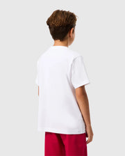 A young person with short hair is standing with their back to the camera. They are wearing a KIDS VINCENT GRAPHIC TEE by Psycho Bunny in plain white, paired with bright red pants against a light gray background.