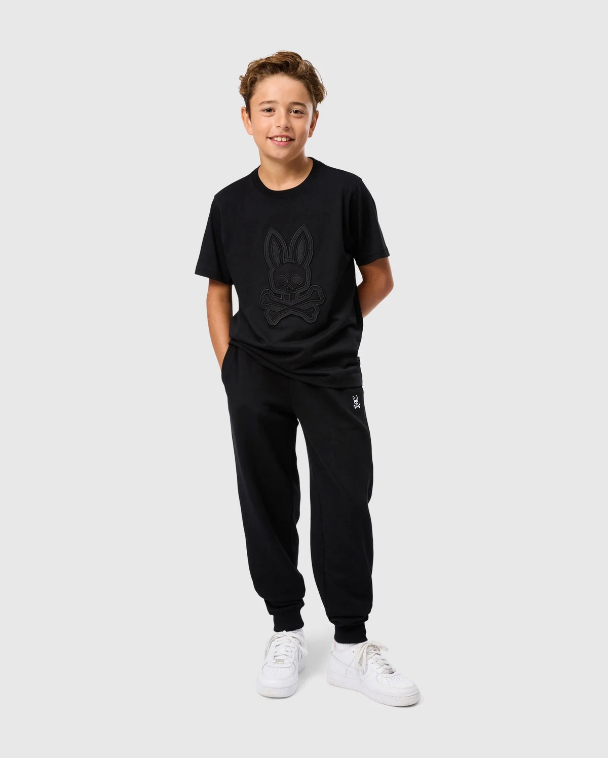 A young boy stands against a plain background, smiling with his hands in his pockets. He is wearing an all-black outfit consisting of the Psycho Bunny KIDS MALTON GRAPHIC TEE - B0U795D200, featuring a cartoon bunny design and matching joggers. The tee showcases Peruvian craftsmanship with its soft Pima cotton fabric. He completes the look with white sneakers.
