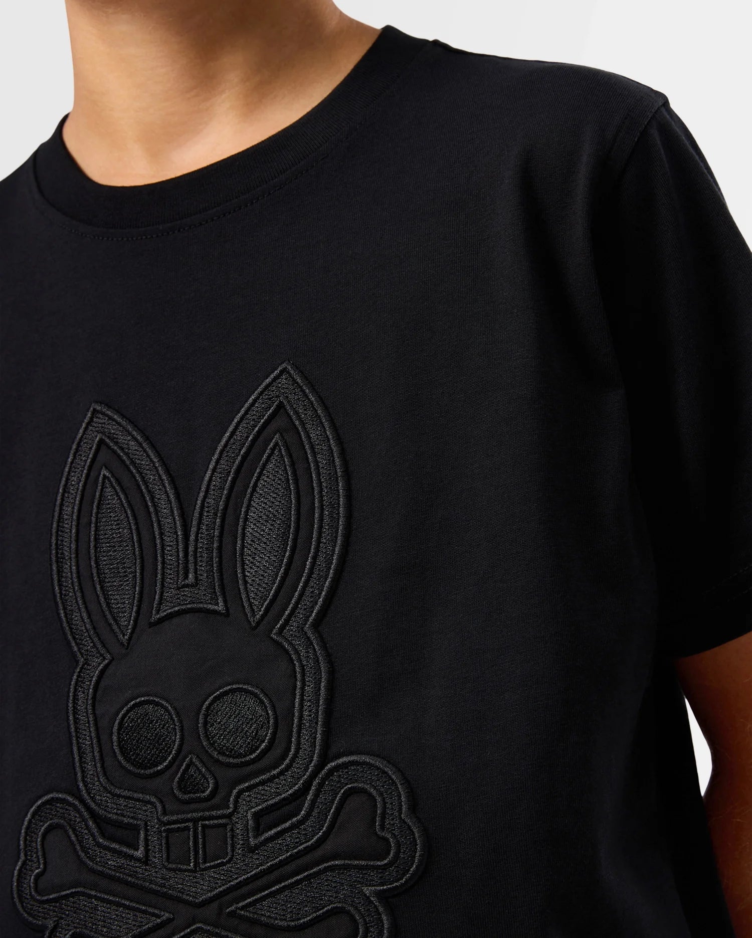 A person wearing a Psycho Bunny KIDS MALTON GRAPHIC TEE - B0U795D200 made from Pima cotton, featuring an embossed design of a skull and crossbones stylized to resemble a rabbit's head with long ears, located on the chest area. The background is light, showing only the upper torso and part of the neck.